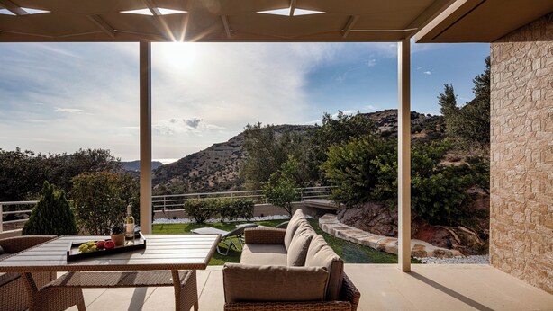 Eine Terrasse im Freien mit Blick auf eine gebirgige Landschaft, ein Tisch mit einer Flasche und Obst sowie ein Korbsessel-Sofa.