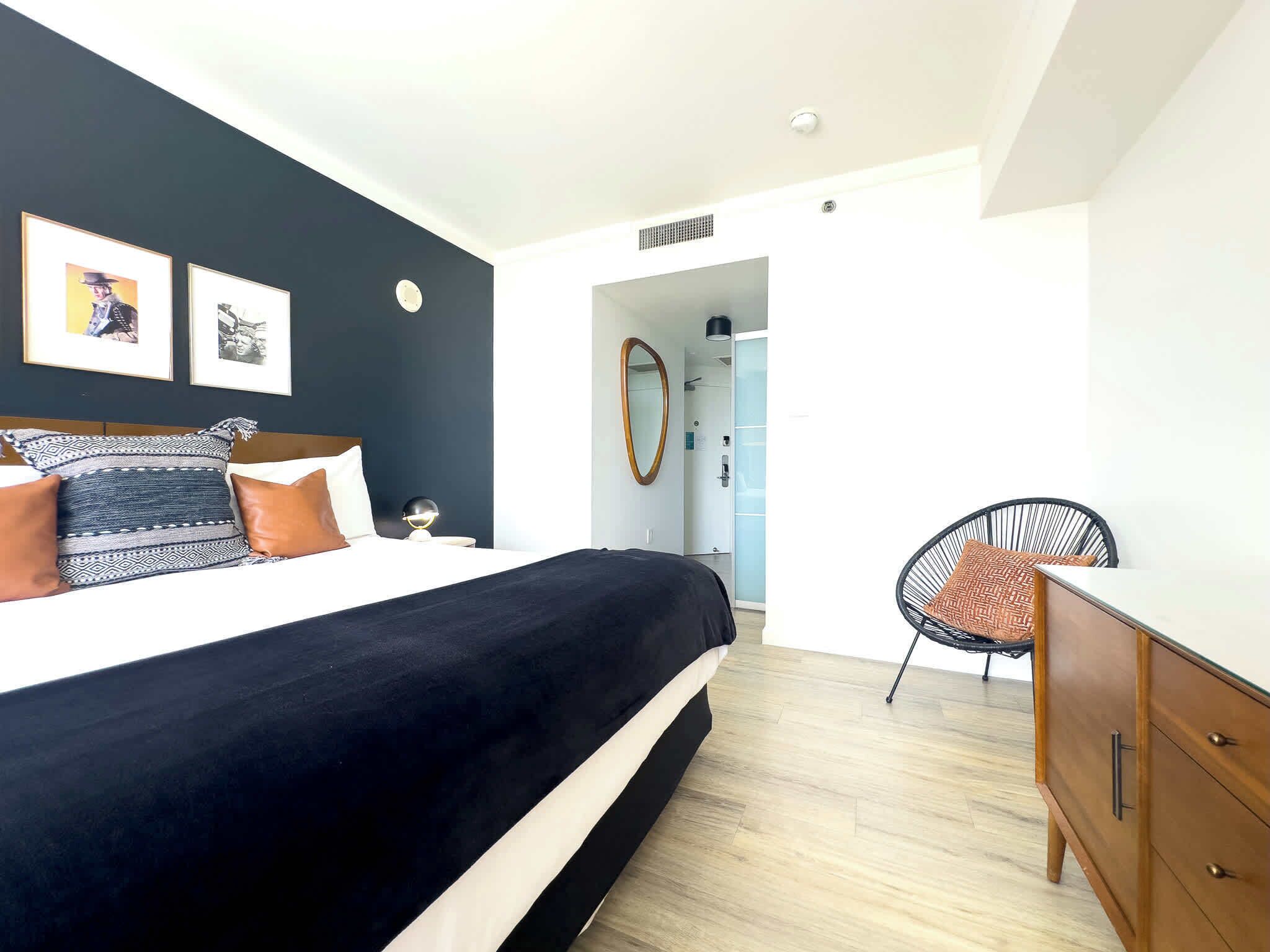 Un dormitorio moderno con una cama grande, un aparador de madera y una silla. Hay cuadros enmarcados en la pared y un espejo sobre el aparador.