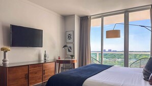 A hotel room with a bed, a TV, a desk with a lamp, and a view of the city.