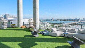 A rooftop terrace with artificial grass, seating, and a view of a city skyline and water.