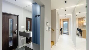 Une chambre d’hôtel moderne avec une porte en bois foncé, un grand miroir, un panneau vertical bleu, un bureau blanc et un coin cuisine comprenant une table et des chaises.