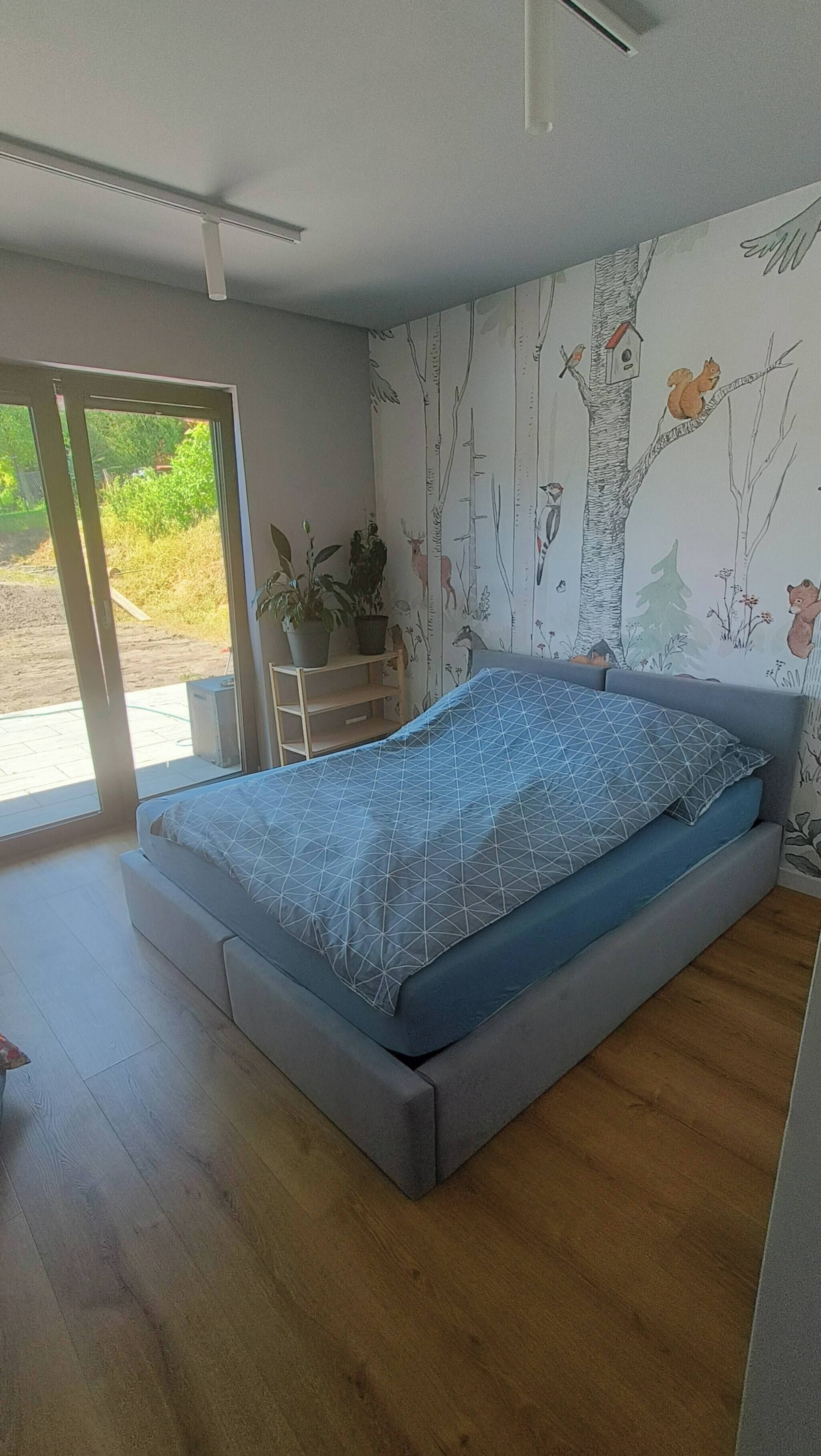 A bedroom with a bed, a window, and a wall with a nature-themed wallpaper.