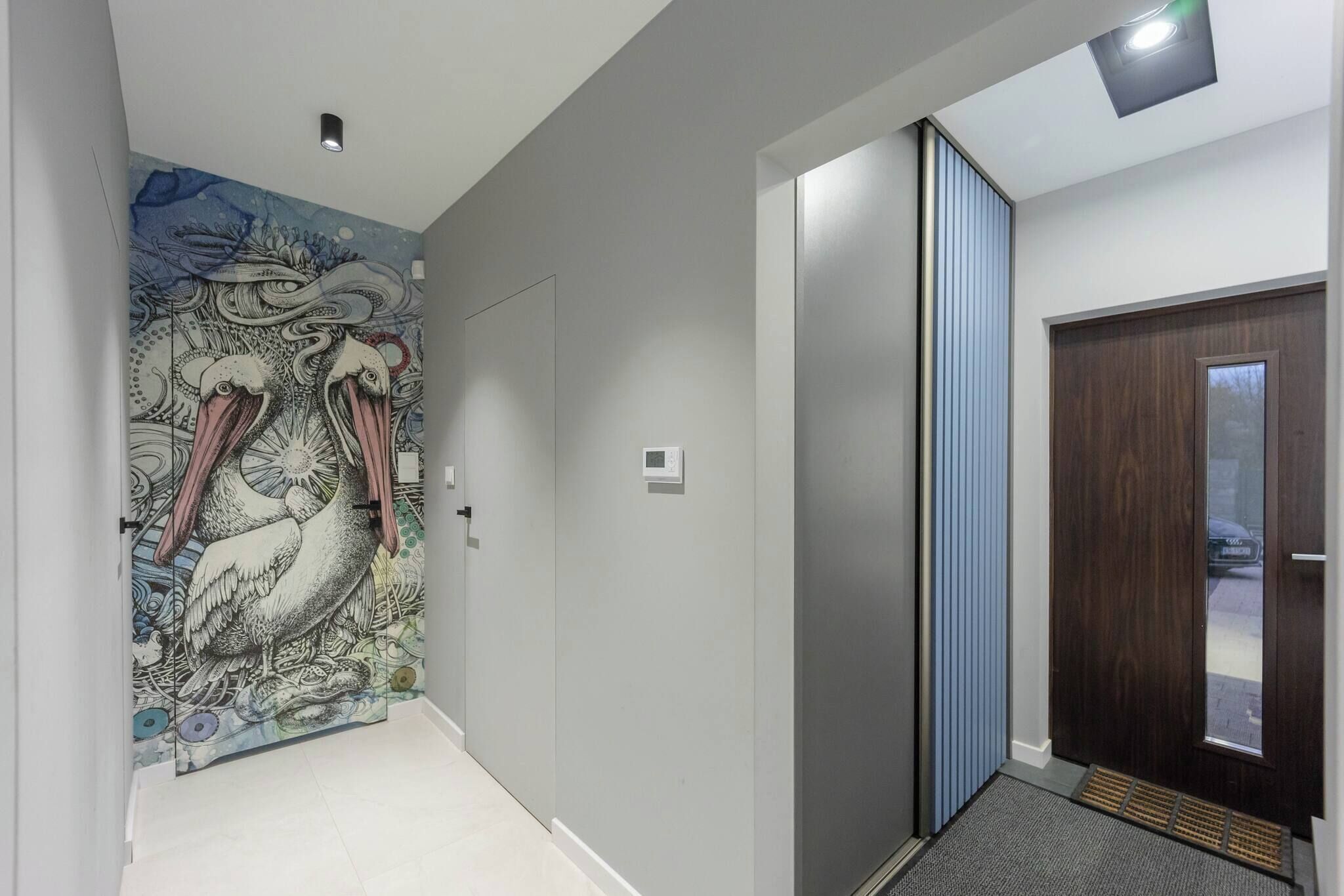 Un pasillo con diseño moderno, que presenta una pared decorativa con un mural de aves, una puerta corredera y una puerta de madera oscura con un panel de vidrio.