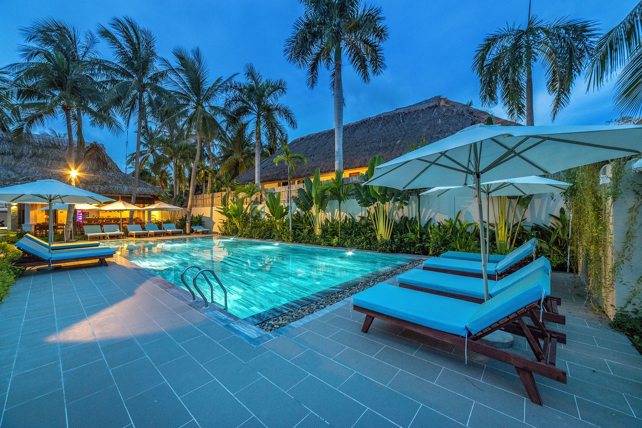 A swimming pool with lounge chairs and umbrellas, surrounded by palm trees and a building with a thatched roof.