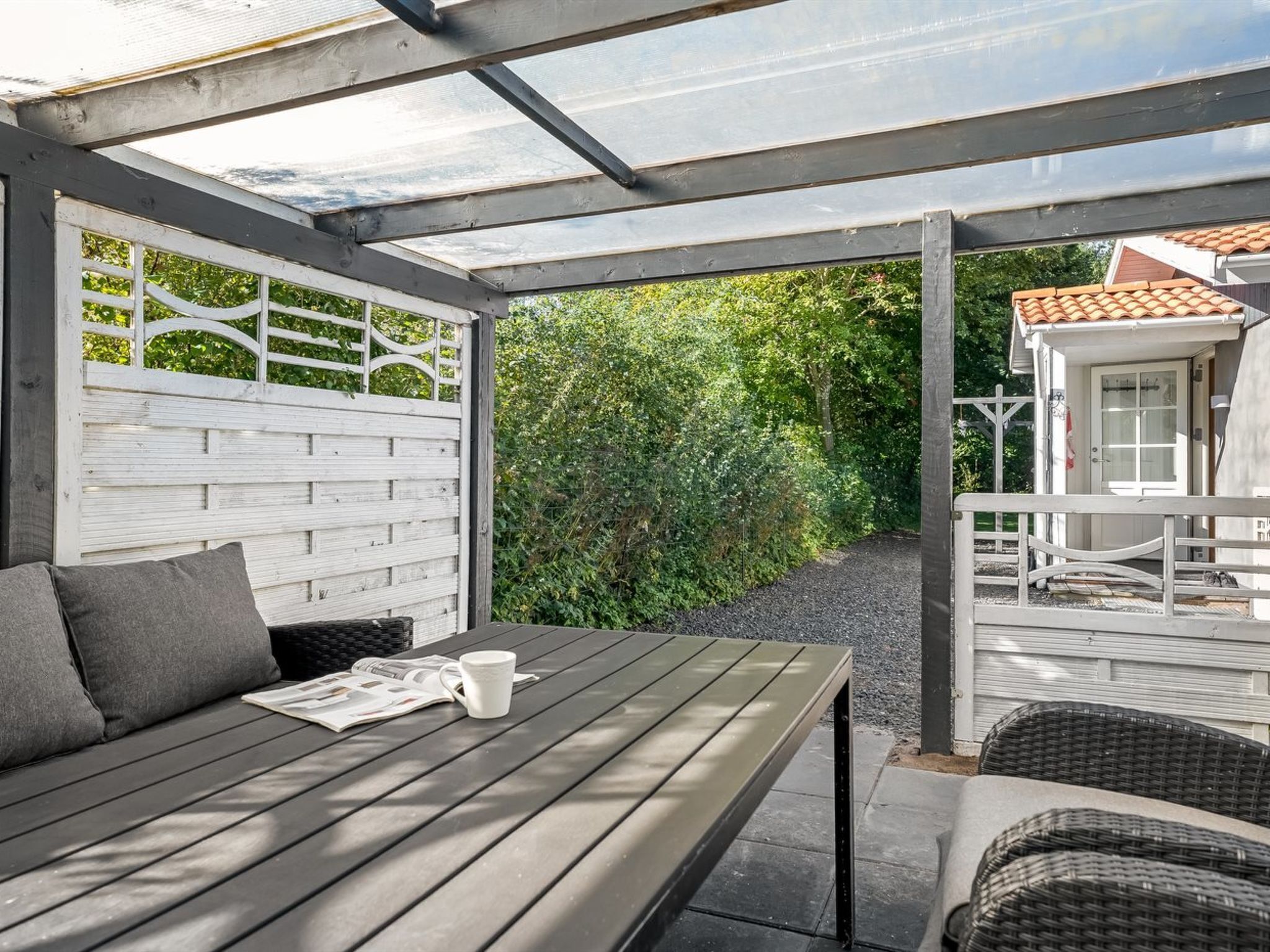 Ein überdachter Patio-Bereich mit einem Holztisch und -stühlen, einem weißen Zaun und Blick auf Grünflächen.