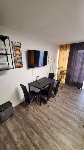 A modern living room with a flat-screen TV, a dining table with chairs, a small shelf with glasses, and a potted plant.