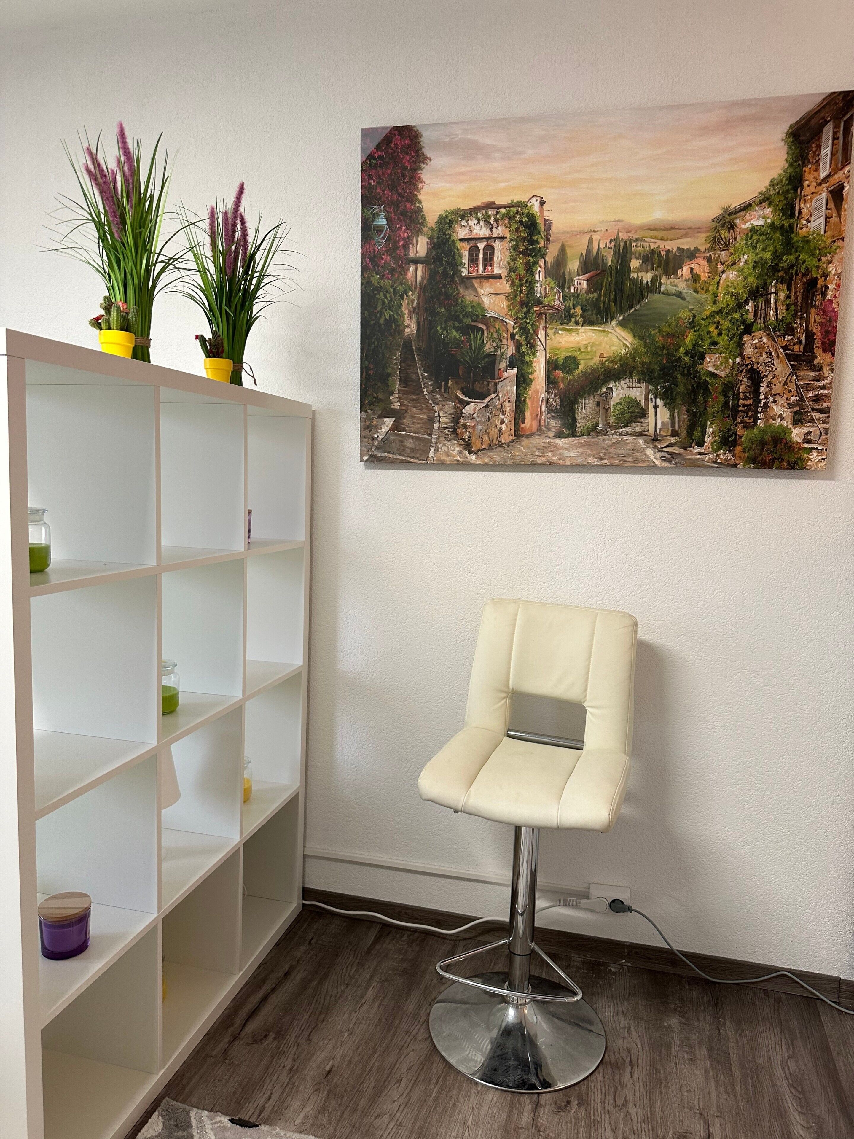 Ein Zimmer mit einem weißen Stuhl, einer weißen Regaleinheit mit Topfpflanzen und einem Landschaftsbild an der Wand.