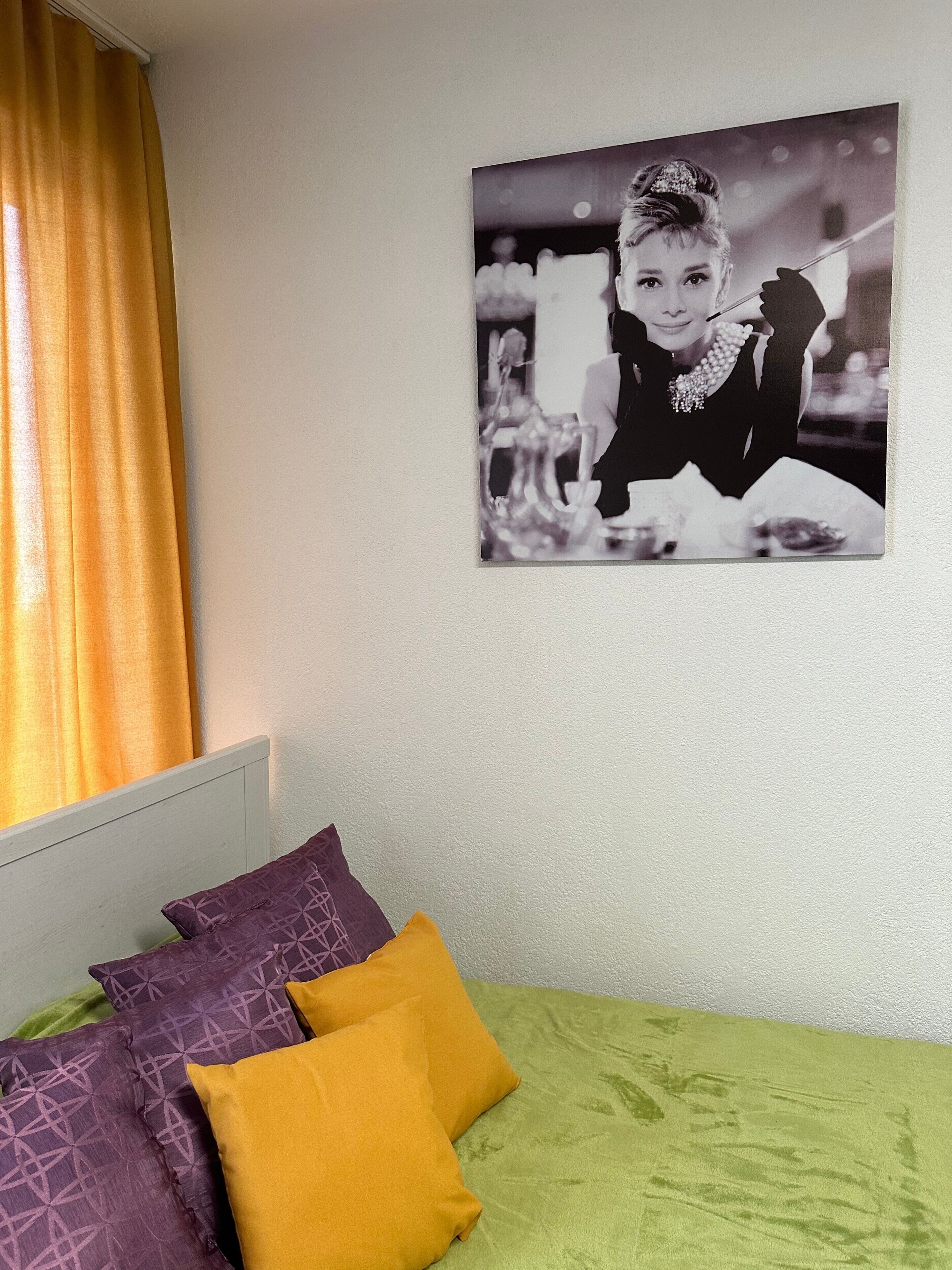 A bed with green and yellow bedding, purple patterned pillows, and a black and white photograph on the wall.