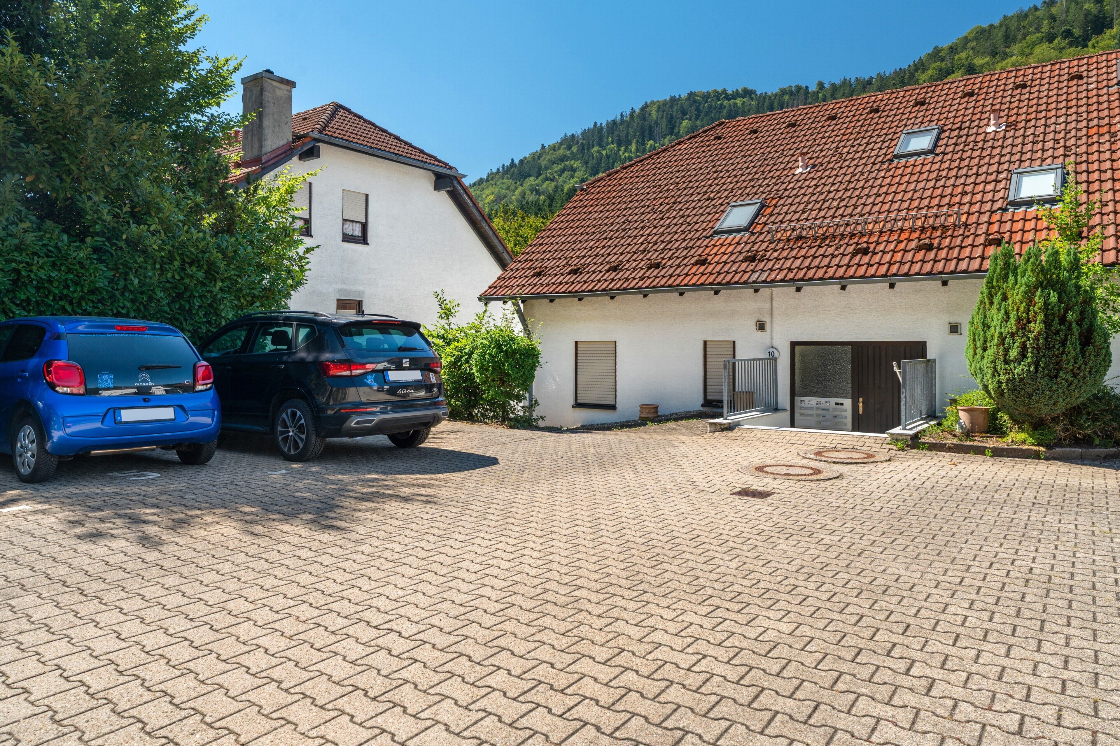 Una strada asfaltata conduce a un edificio bianco con tetto in tegole rosse, auto parcheggiate e montagne sullo sfondo.