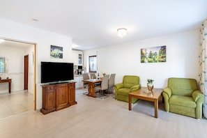 A living room with a TV, green armchairs, a wooden coffee table, and a dining area with chairs and a table.