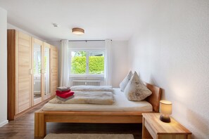 A bedroom with a wooden bed, a bedside table with a lamp, a window with a view of greenery, and a wardrobe.