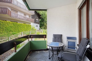 A balcony with a table and chairs, a view of a building, and a green hedge.