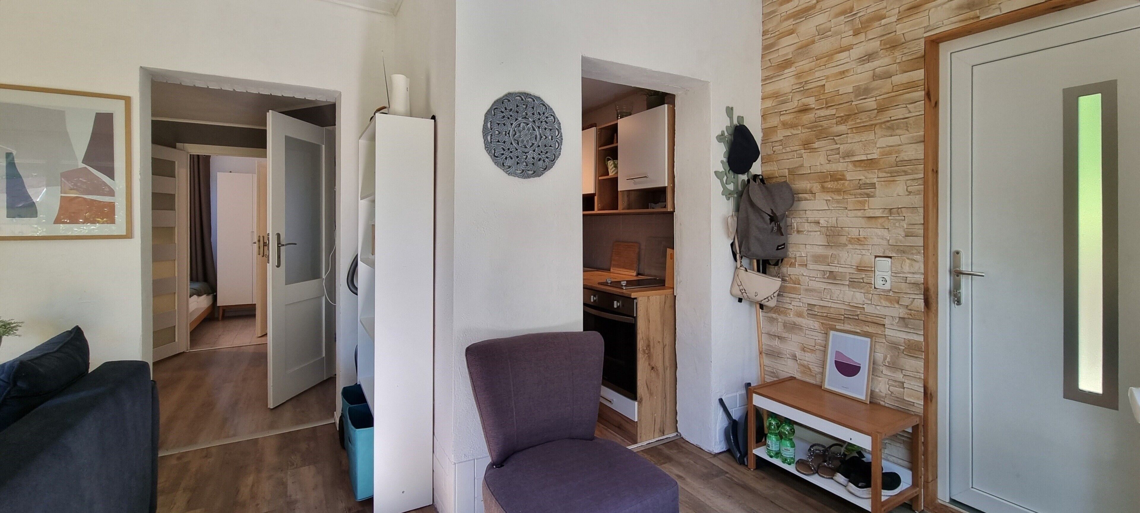 A living room with a purple armchair, a white refrigerator, a kitchen with wooden cabinets, and a stone wall.