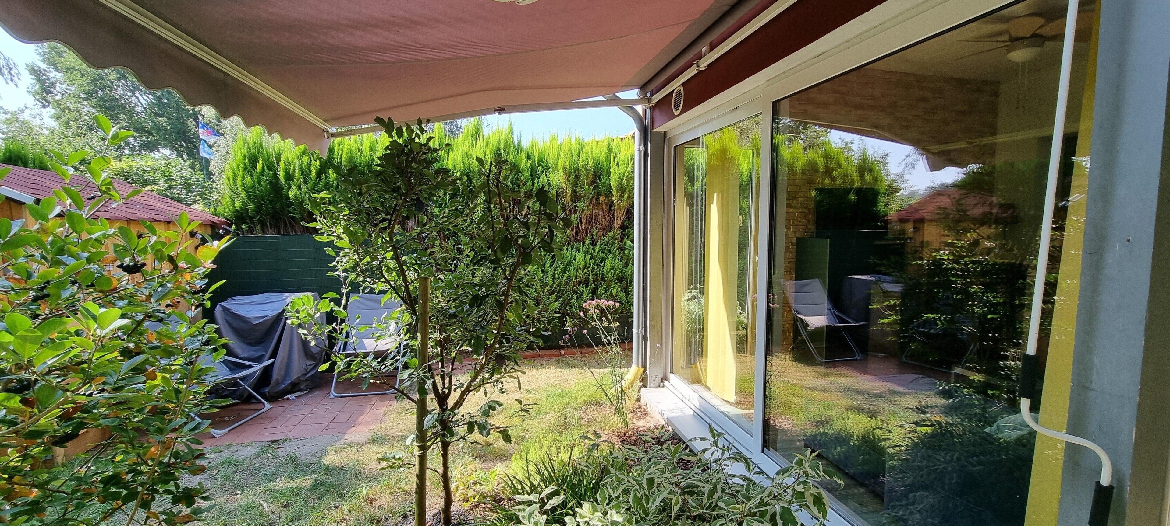 A covered patio with a table and chairs, a small tree, and a glass door leading to a garden.