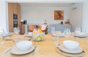 Un coin repas avec une table en bois mise pour le repas, un panier de fruits et, au fond, une cuisine.