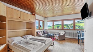 A bedroom with a bed, a sofa, a chair, a small table, and a view of a green landscape through large windows. - Haus Kehrwieder Studio with Panoramic Views, Hochschwarzwald Card, Wi-Fi, Parking (Todtnau)