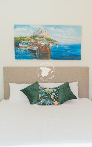 A bed with white linens and two green pillows, a beige headboard, and a painting of a coastal scene above the bed. - CostaBlancaDreams - Casa Monica in Calpe (Calpe)