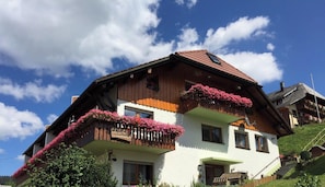 Un chalet Ă deux Ă©tages dotĂ© de balcons en bois ornĂ©s de fleurs, dâun toit en pente et surplombant un ciel dĂ©gagĂ©.