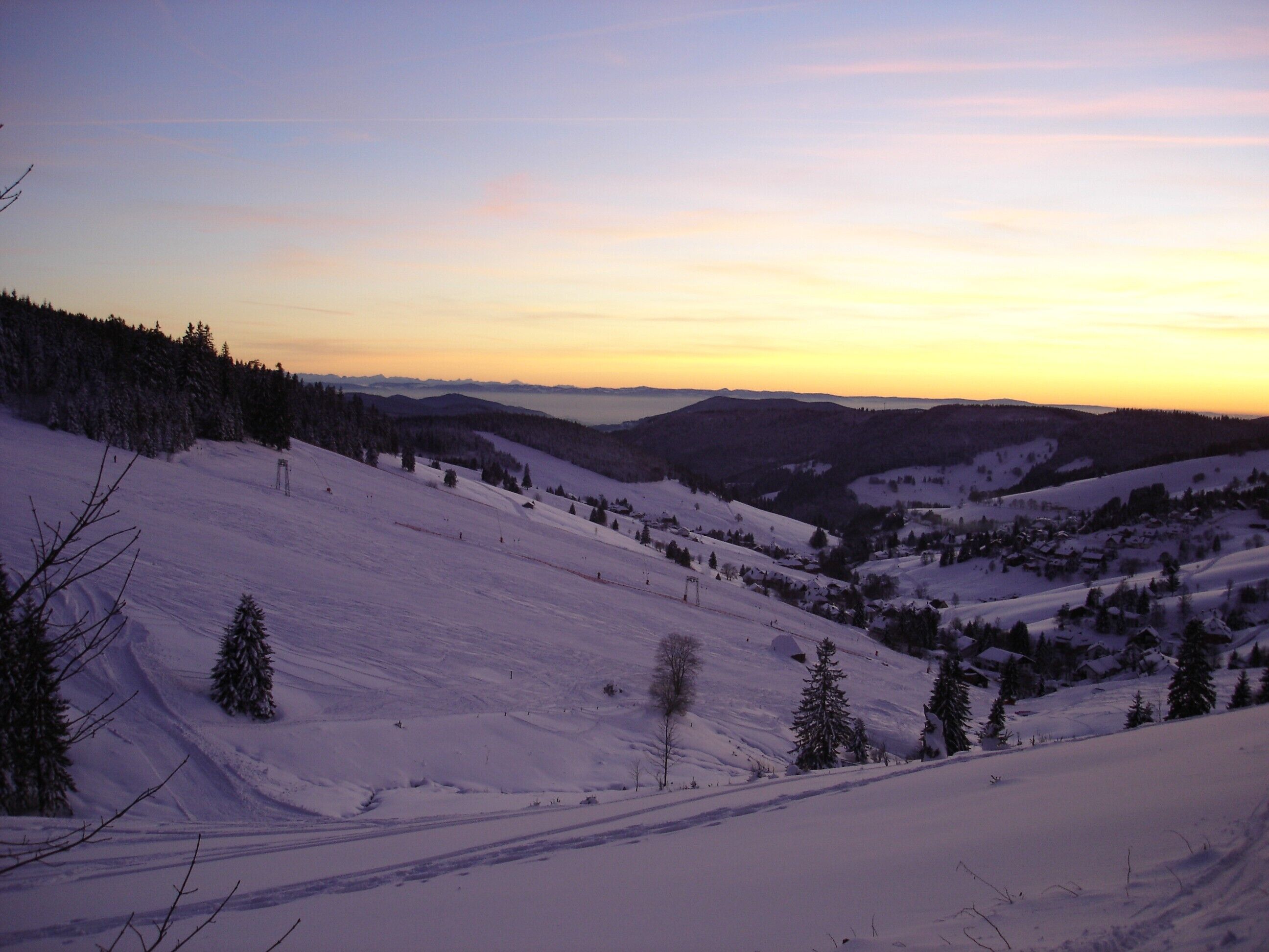 A snowy landscape with a clear sky at sunset, featuring a valley with scattered trees and distant hills.