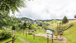 Un parque infantil con tobogán y estructuras para escalar, rodeado de vegetación y vistas a un pueblo lejano.