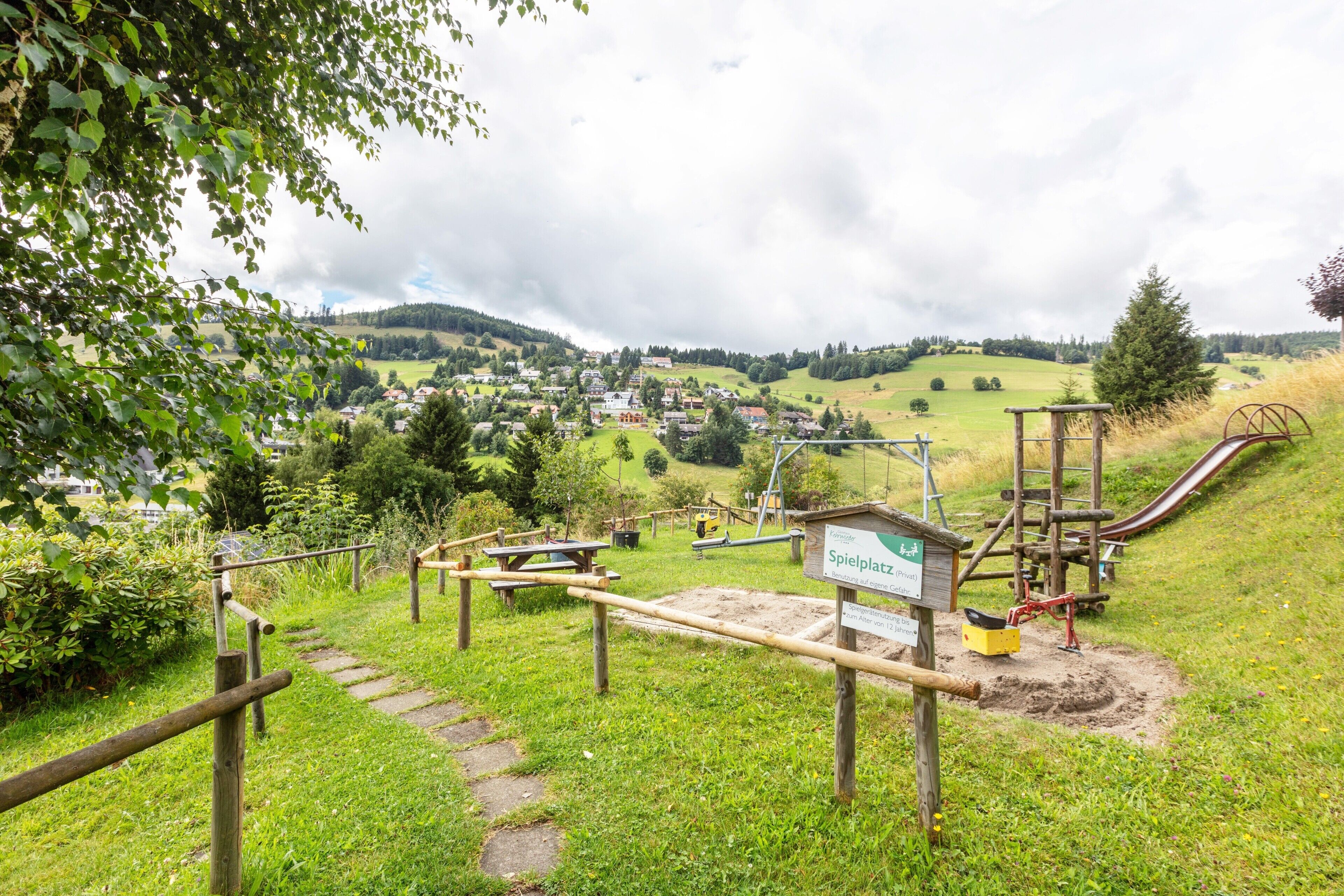 Ein Spielplatz mit Rutsche und Klettergerüsten, umgeben von Grünflächen und Blick auf ein Dorf in der Ferne.