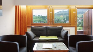 Een kamer met een bank, een tafel met een afstandsbediening, en uitzicht op een groen landschap door grote ramen.