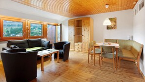 Een kamer met een houten plafond, een bank, een tafel, stoelen, een houten kast en een raam met uitzicht op groen.