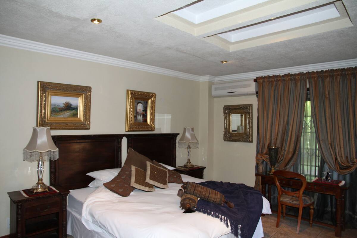 A hotel room with a large bed, two bedside tables with lamps, a wooden chair, and a desk with a chair. There are two framed pictures on the wall and a window with curtains.