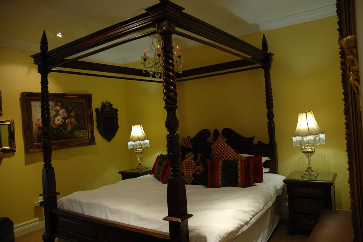 A four-poster bed with a wooden frame and a canopy, two bedside tables with lamps, a painting on the wall, and a mirror.