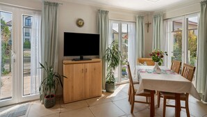 Une pièce avec une télévision, un meuble en bois, une table à manger, des chaises, des plantes en pot et de grandes fenêtres avec des rideaux.