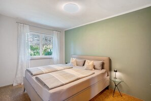 A bedroom with a bed, a bedside table, a window with curtains, and a view of greenery outside.