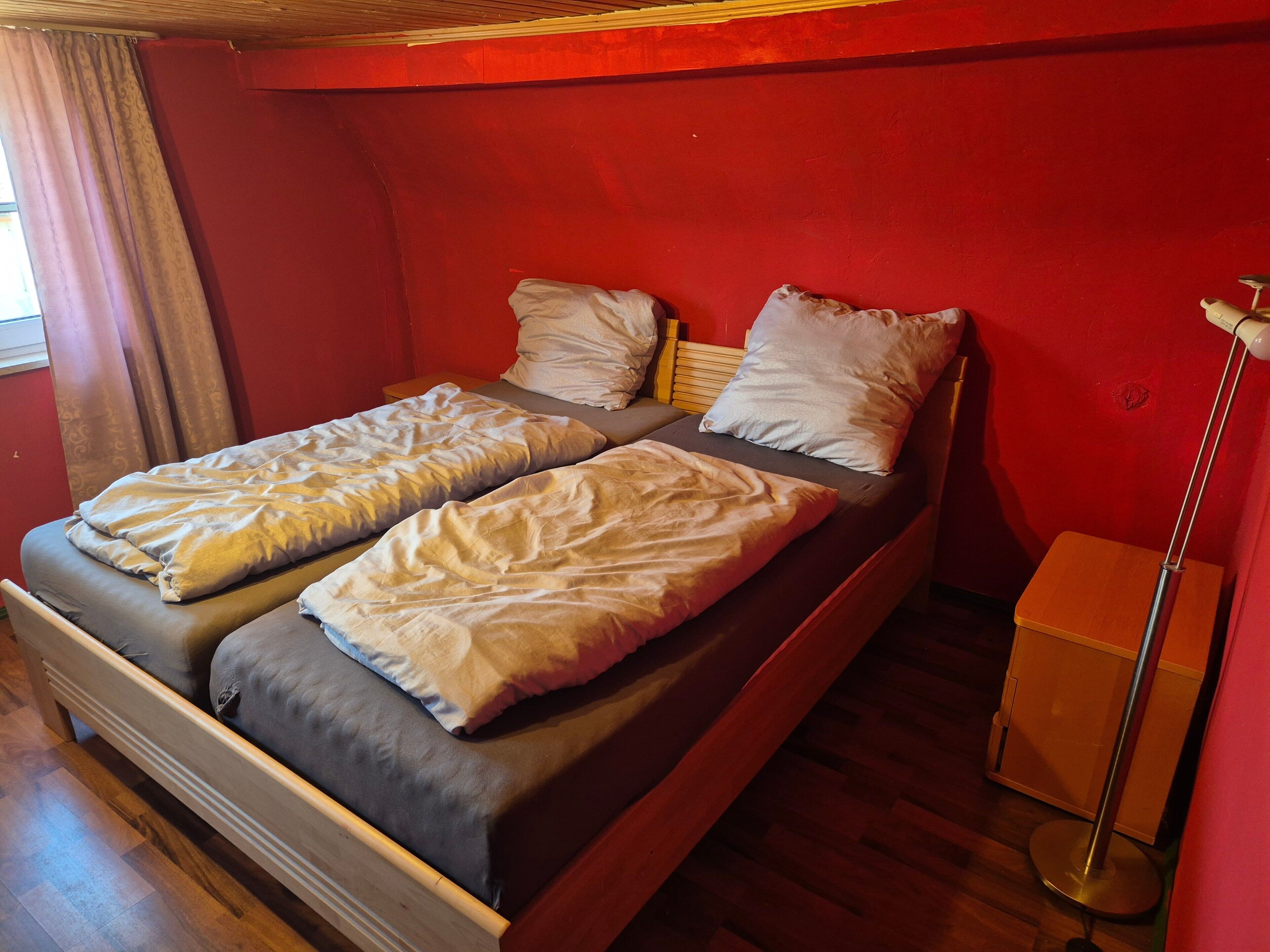 Uma cama de casal com duas almofadas, uma mesinha de cabeceira e um abajur em quarto com paredes vermelhas e piso de madeira.