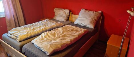 Uma cama de casal com duas almofadas, uma mesinha de cabeceira e um abajur em quarto com paredes vermelhas e piso de madeira.
