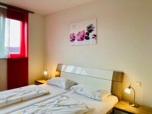 Una habitación de hotel con una cama, dos mesitas de noche, un ventanal con cortinas rojas y una obra de arte colgada en la pared.