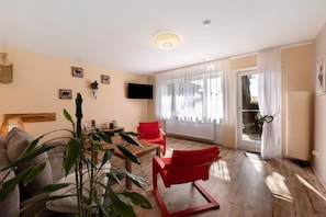 Un salon avec un canapé, un fauteuil rouge, un téléviseur fixé au mur et une fenêtre avec des rideaux.