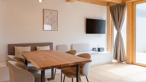 A modern living room with a wooden dining table, cushioned seating, a flat-screen TV, and a large window with curtains.