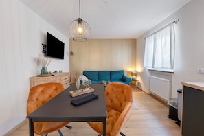 A modern living room with a sofa, a dining table with chairs, a TV mounted on the wall, and a window with curtains.