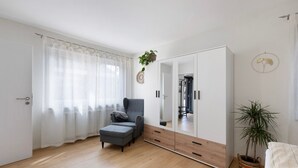 A room with a large wardrobe, a grey armchair, a small ottoman, a potted plant, and a bed with a striped blanket.