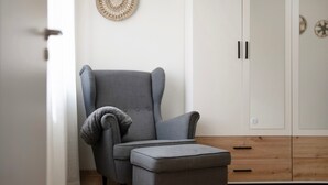A room with a grey upholstered armchair, a matching ottoman, a wooden cabinet with drawers, and a woven wall decoration.