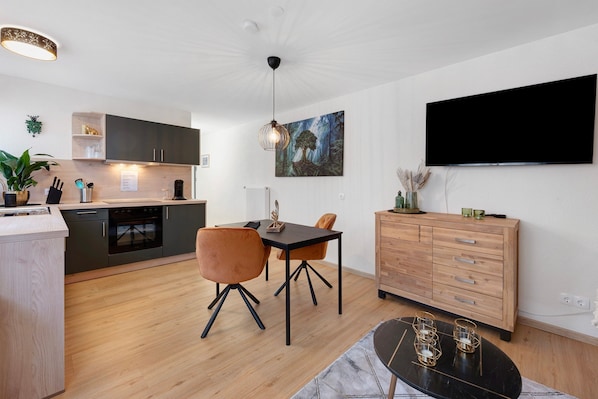 A modern kitchen with dark cabinets, a dining table with chairs, a black TV mounted on the wall, and a wooden cabinet with drawers.