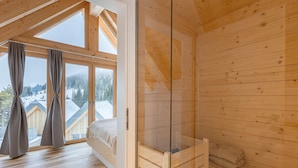Quarto de cabana de madeira com cama, box de vidro para o chuveiro e grandes janelas com vista para uma paisagem nevada.
