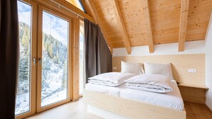 A bedroom with a wooden ceiling, a bed with white bedding, and a view of a snowy landscape through large windows.