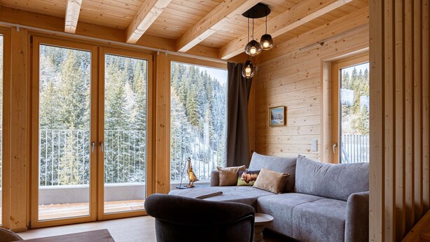 A wooden cabin interior with a sofa, a chair, and a table. There are large windows with a view of trees.