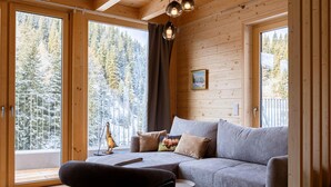 A cozy wooden cabin interior with a grey sofa, a small round table, and a view of snow-covered trees through large windows.