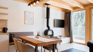 A cozy room with a wooden table, chairs, a sofa, a fireplace, a TV, and a large window with a view of trees.
