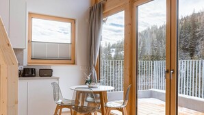 A cozy room with a wooden table and chairs, a small kitchen area, and a balcony with a view of a snowy forest.
