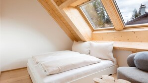 A small, cozy room with a sloped ceiling, a bed, a chair, and a window with a view of trees and buildings.