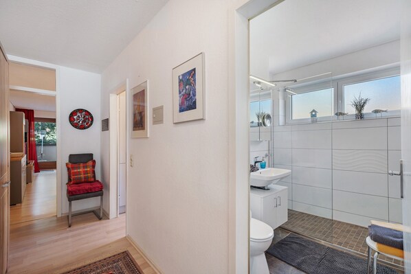 Ein Flur, der zu einem Badezimmer mit Waschbecken, Toilette und Fenster führt. Eine Bank mit roter und grüner Decke, ein gerahmtes Bild an der Wand und eine dekorative Platte sind ebenfalls vorhanden.