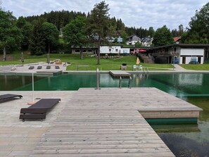 A wooden dock leading into a swimming pool with a grassy area and houses in the background. - Apartment '101 Luisa' with Mountain View, Balcony and Wi-Fi (Schönwald im Schwarzwald)