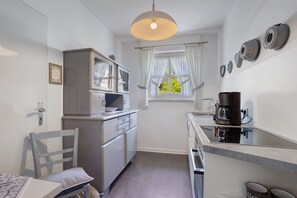 A compact kitchen with a refrigerator, stove, and coffee maker. A window with curtains provides natural light. - Apartment 'Ferienwohnung Riedel' with Balcony and Wi-Fi (Sundern (Sauerland))